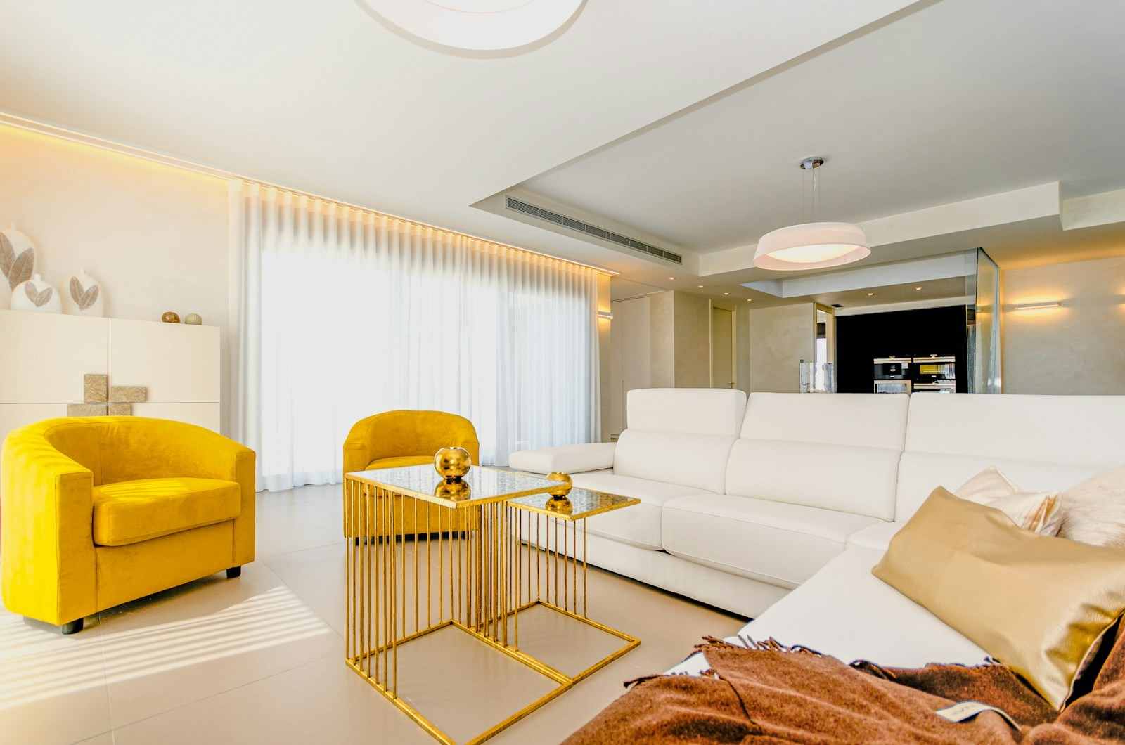 Upscale San Francisco apartment living room featuring a long white leather sectional sofa, two striking yellow velvet accent chairs, and contemporary gold nesting side tables. This bright interior design showcases premium finishes, layered lighting, and sheer white drapery, perfect for high-end properties in the 94110 zip code area of the Mission District. This stunning, brightly lit living space emphasizes contemporary luxury ideal for modern San Francisco homes. The design centers around a comfortable white leather sectional, providing a neutral anchor, while two matching yellow velvet armchairs introduce a sophisticated pop of color and texture. Gold metal nesting side tables add a layer of high-end elegance, complementing the clean white walls and sheer drapery that maximize natural light in this expertly styled interior.
