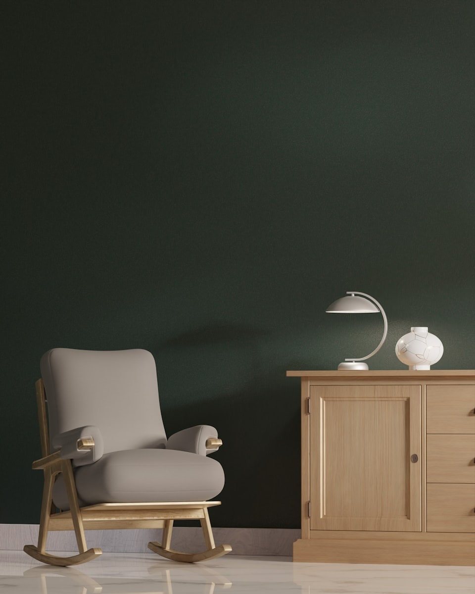Modern interior scene featuring a light beige upholstered rocking chair with a natural wood frame, positioned against a dark forest green accent wall, next to a wood sideboard decorated with a minimalist white lamp, perfect for cozy reading in a San Francisco home. This sophisticated interior rendering showcases how deep, moody colors, like the dark green accent wall, can create a cozy yet modern atmosphere. The soft taupe rocking chair provides a comfortable retreat, balanced by the warmth of the natural wood cabinet. This design approach is highly sought after for luxury urban apartments and homes in areas like the Mission District of San Francisco, offering an elegant contrast essential for chic Bay Area living spaces.