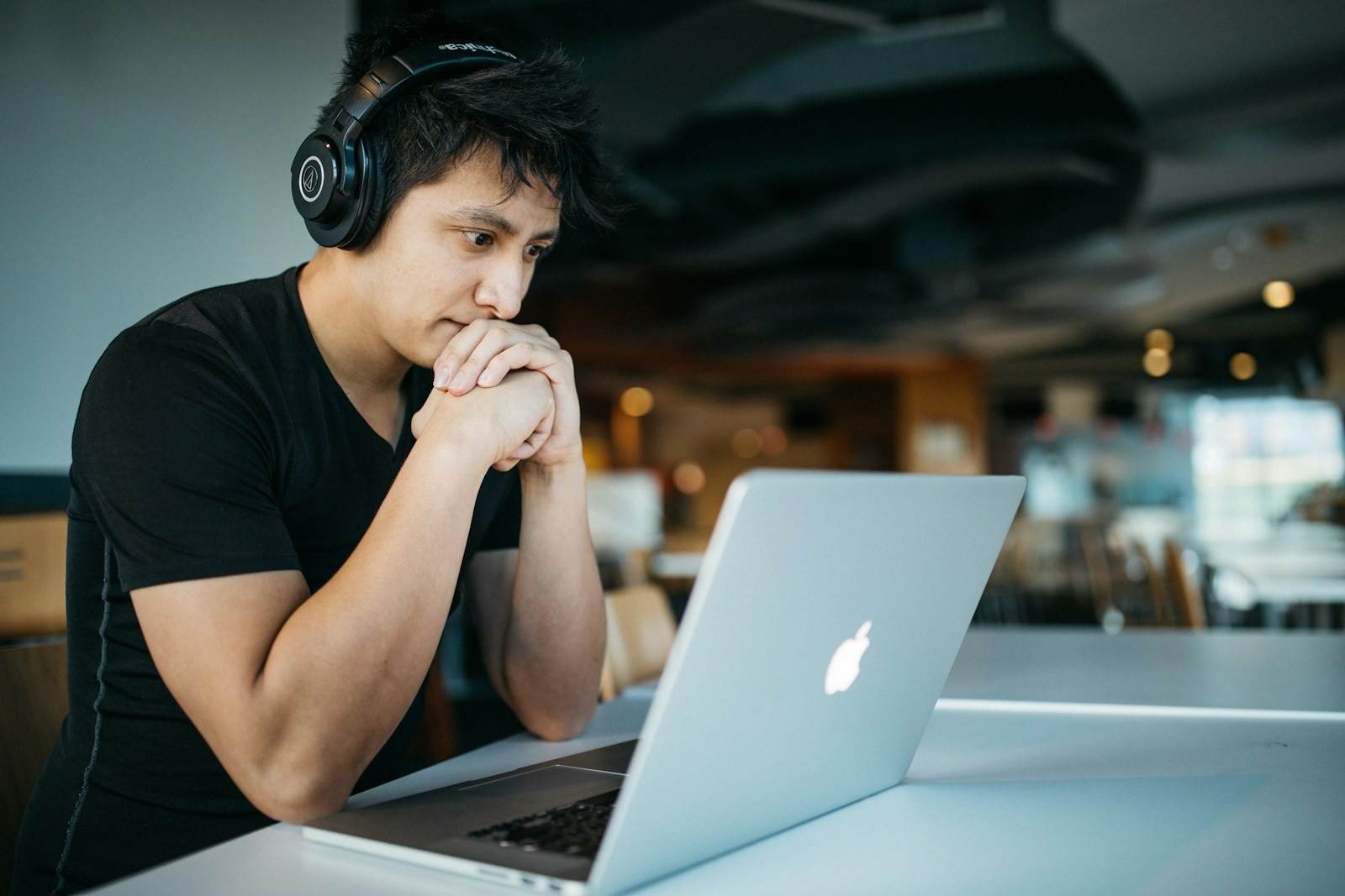Young Asian man with black headphones intently focused on a silver MacBook laptop while working remotely in a bright, modern San Francisco co-working space or cafe setting. This image captures the dedication of modern professionals utilizing the flexibility of remote work. A young man, wearing noise-canceling headphones, pores over his laptop screen, symbolizing the focused effort required for coding, studying, or virtual collaboration. The setting reflects the dynamic, tech-savvy environment prevalent in the San Francisco Bay Area, where productivity often happens outside traditional office walls.