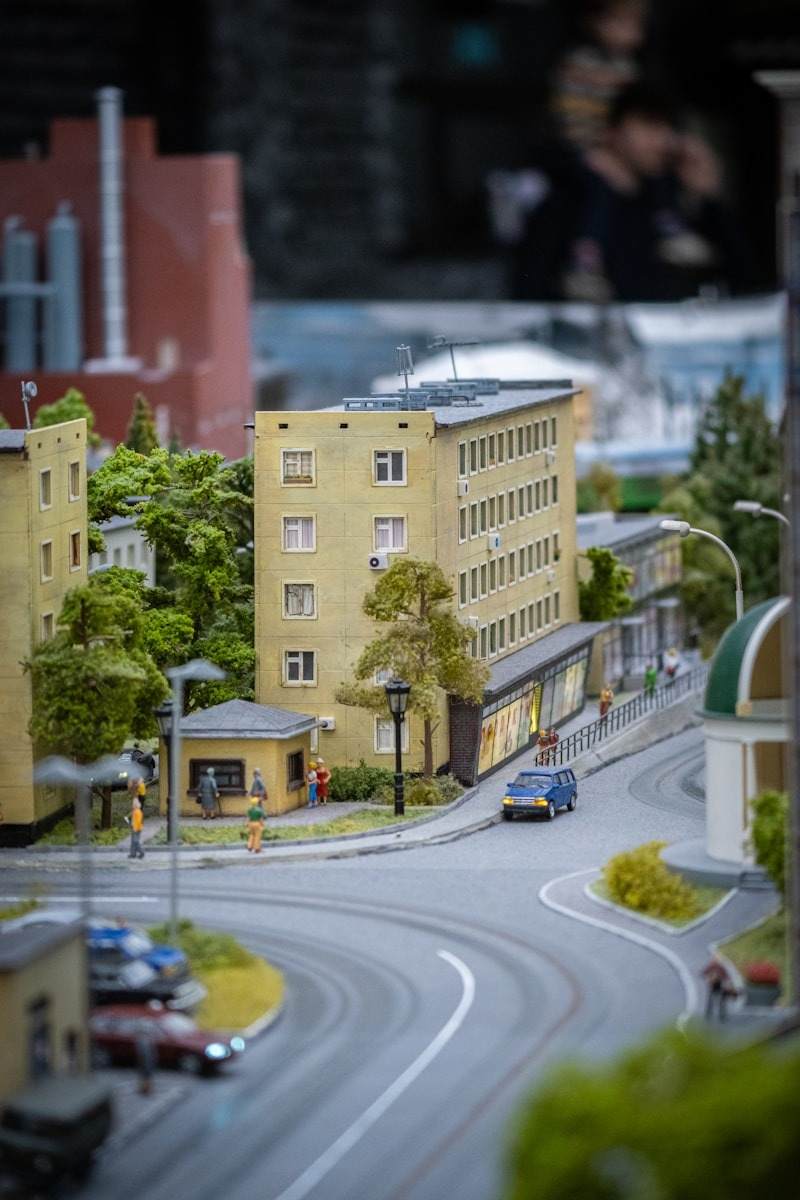 Highly detailed miniature cityscape diorama featuring yellow residential buildings, lush green trees, streetlights, and tiny model cars driving on a winding urban road, often seen in sophisticated model art exhibits near the Mission District, San Francisco. Explore the meticulous detail captured in this urban miniature diorama, showcasing yellow apartment buildings and realistic street environments. This type of intricate scale modeling demonstrates superior craftsmanship, echoing the complex architectural density found in vibrant neighborhoods like the Mission District of San Francisco. Such exhibits provide a unique perspective on urban planning and artistic representation, appealing to hobbyists and art enthusiasts exploring the city's cultural offerings.