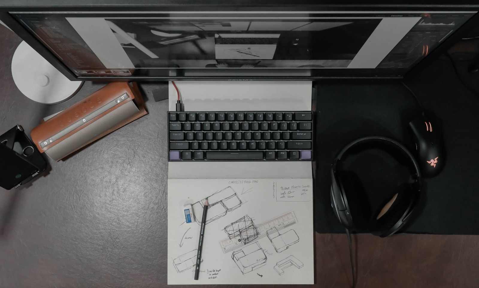Top-down view of a focused creator's desk setup featuring a compact black mechanical keyboard, Razer mouse, black over-ear headphones, a monitor, and a notebook showing detailed product design sketches near Mission District, San Francisco. This overhead shot captures the essential tools of a modern digital and product designer. Centered around a sleek mechanical keyboard, the desk also holds a monitor displaying design work, high-quality headphones, and a notebook filled with preliminary technical drawings (including product concepts labeled 'CANDLES PARA MAN'). This mix of high-tech gear and traditional sketching tools exemplifies the creative workflow often found among the innovative professionals operating near the Mission District in San Francisco, emphasizing both focused execution and conceptual development.