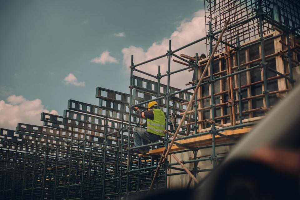 A dedicated construction worker wearing a yellow hard hat and high-visibility vest sits on wooden scaffolding planks, securing massive metal formwork panels for a multi-story concrete structure under a cloudy sky in San Francisco's Mission District. This photograph captures the intensity of urban development, showing a skilled construction worker perched on intricate metal scaffolding. The worker, wearing essential safety gear including a hard hat and high-visibility vest, is actively involved in setting up the concrete formwork for a significant new structure near the Mission District of San Francisco. This represents the ongoing commitment to infrastructure growth, illustrating the careful planning and complex engineering challenges faced by Bay Area builders.