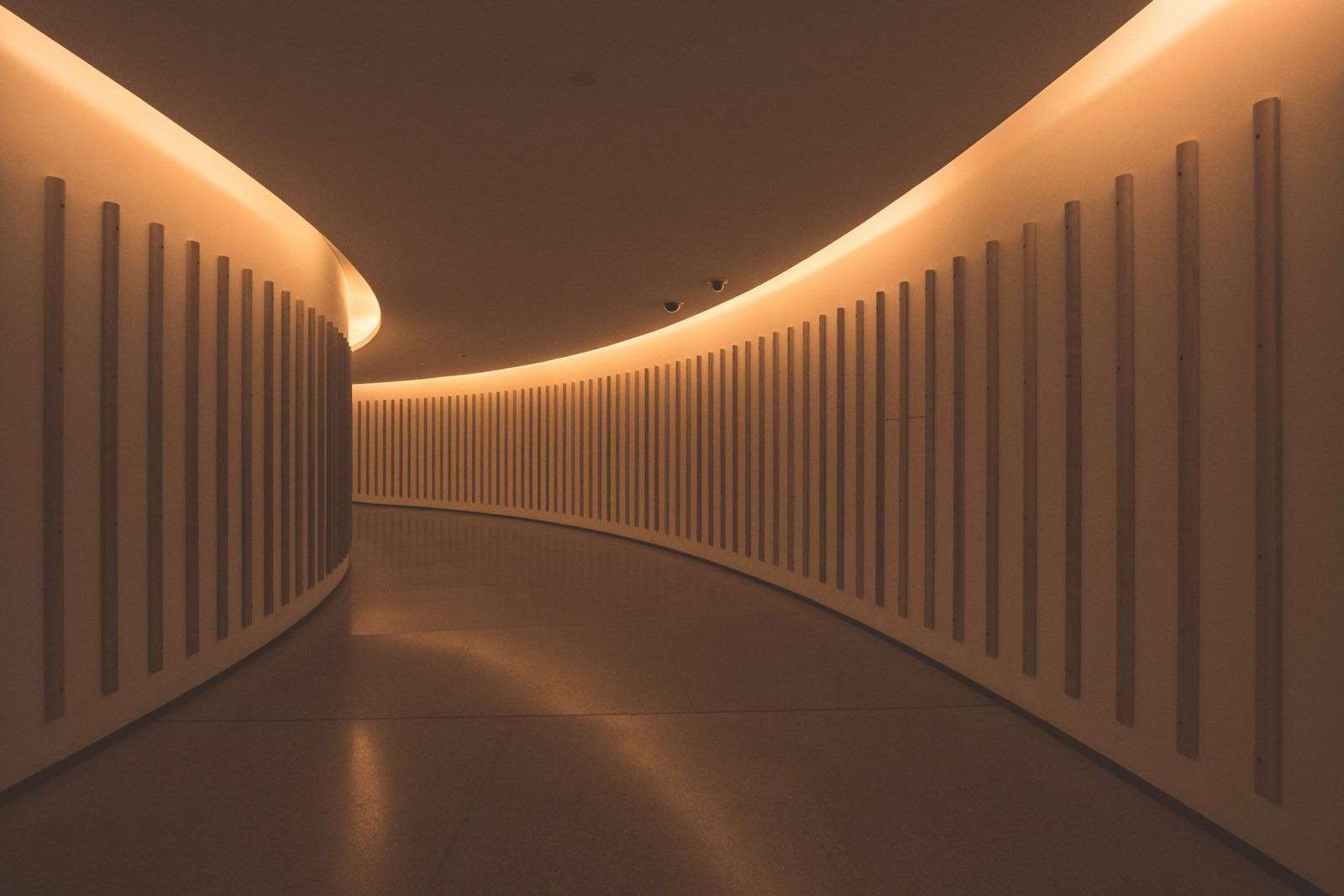 Architecturally stunning curved corridor featuring vertical paneling and warm amber cove lighting, illustrating modern interior design elements inside a commercial building in San Francisco. This photograph captures a striking example of contemporary interior architecture, featuring a gently curving hallway illuminated by soft, continuous amber cove lighting. The minimalist aesthetic is enhanced by the repeating pattern of vertical slats against the smooth walls. This design evokes the sophisticated, modern spaces often found throughout the vibrant arts and commercial districts of San Francisco, offering inspiration for innovative spatial planning and architectural lighting solutions in the local area.