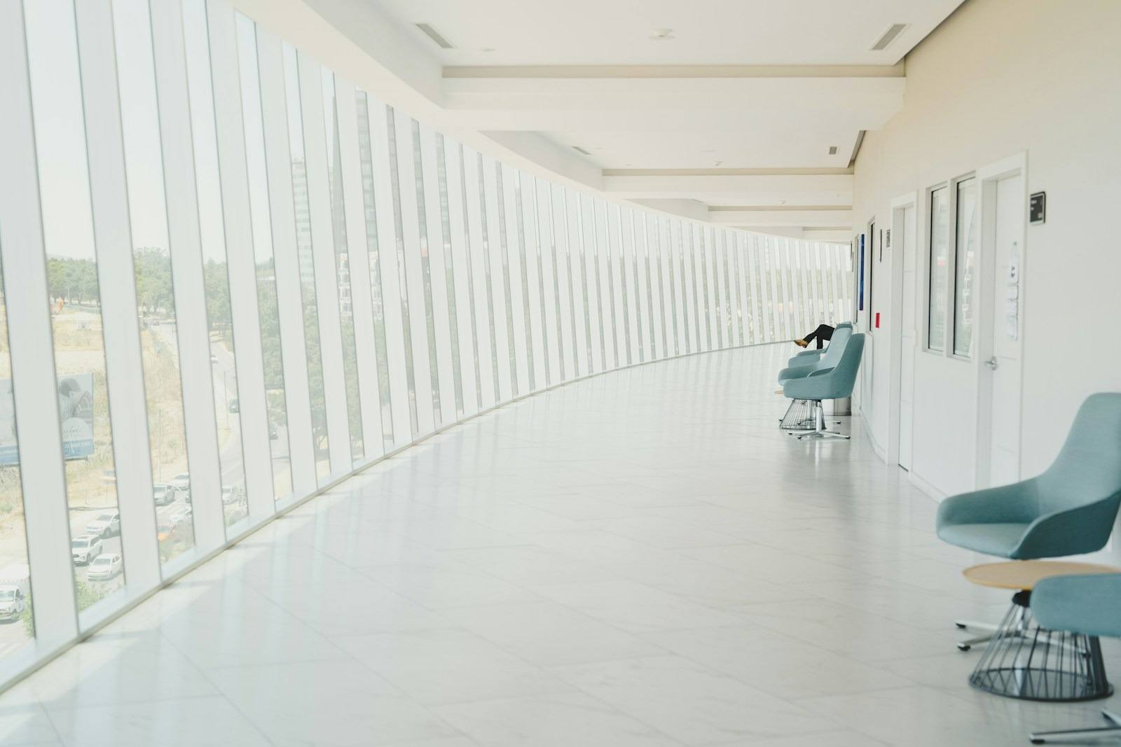 Bright, modern, curved architectural hallway in a San Francisco commercial or medical building waiting area, featuring light teal swivel chairs, white tile floors, and floor-to-ceiling windows providing natural light and city views near Lat 37.76, Lng -122.41. This photograph captures the serene and expansive design of a modern corporate or healthcare building hallway, characteristic of recent San Francisco commercial architecture. The bright, curved space is lined with tall, vertical windows, maximizing natural light and offering a dynamic perspective of the exterior urban landscape. Minimalist teal swivel seating provides comfortable waiting spots, reinforcing an environment that is both professional and calming for visitors in the Bay Area.