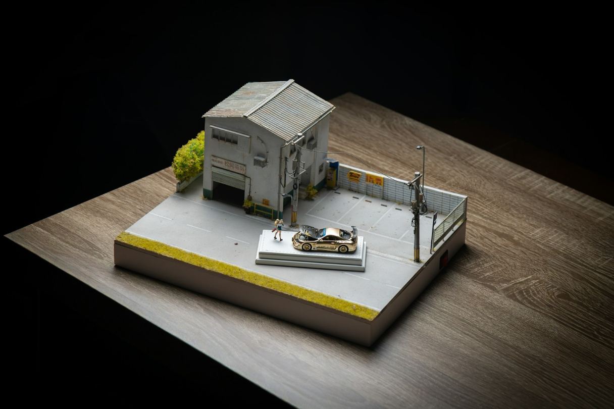 Highly detailed miniature diorama featuring a metallic gold JDM tuner car with an open hood, parked on a street platform next to a weathered Japanese-style garage, showcasing intricate model making artistry popular among collectors in the San Francisco Mission District. This stunning photograph captures a handcrafted scale model diorama dedicated to automotive culture and intricate miniature architecture. The scene depicts a customized gold JDM sports car displayed outside a realistic, worn-down repair shop, complete with detailed surroundings like fencing, power lines, and a miniature figure. Such fine art scale modeling resonates strongly with collectors and creators, mirroring the appreciation for precision engineering and detailed craftsmanship found throughout the Bay Area, especially among hobbyist communities located near Mission Dolores Park in San Francisco.