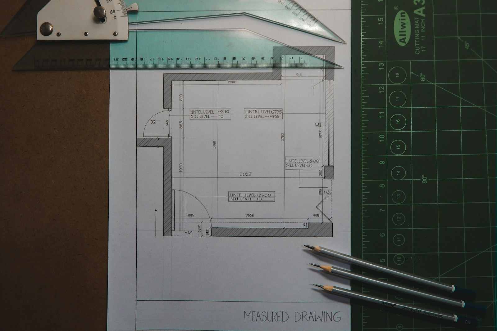 Detailed hand-drawn architectural measured drawing and floor plan, showing metric dimensions and lintel/sill levels, resting on an A3 cutting mat with drafting tools and pencils, symbolizing precise architectural surveying and design work for remodeling projects in the Mission District, San Francisco (37.76 N, 122.41 W). Accuracy is paramount in architectural design, particularly in complex urban environments like San Francisco. This image captures a moment of meticulous preparation, showcasing a hand-drafted measured drawing complete with precise metric dimensions, door, and window notations (D1, D2, W1) and structural levels (lintel and sill). Such detailed surveying work, often performed by skilled technicians or architects, is crucial for ensuring that renovations or new construction projects adhere perfectly to existing structures and local planning codes throughout the Bay Area. This process underpins successful project execution for architecture firms operating near 37.76 N, 122.41 W.
