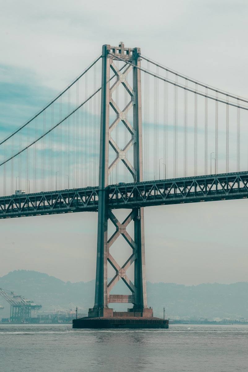 Vertical photograph of the central tower pier and steel suspension structure of the San Francisco-Oakland Bay Bridge rising from the water, visible from the San Francisco side of the Bay. This close-up view captures the massive foundational pier and the architecturally distinctive lattice tower of the San Francisco-Oakland Bay Bridge. The bridge's steelwork and intricate cables dominate the frame, rising impressively from the waters of the San Francisco Bay. This infrastructure is crucial, linking the city of San Francisco (near 37.767 N, -122.419 W) with the East Bay, symbolizing regional commerce and transportation against a backdrop of distant hills and port activity.