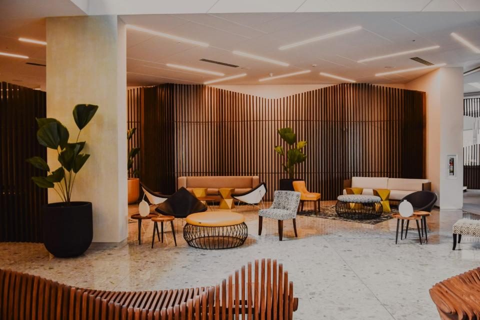 Modern luxury lounge interior in San Francisco near Civic Center (37.767296, -122.419498), featuring contemporary seating arrangements, a prominent dark wood vertical slat accent wall, large potted indoor plants, and polished light terrazzo flooring. This inviting and contemporary common area is representative of high-end San Francisco interior design near the Civic Center area. The space utilizes rich materials, including light terrazzo flooring and a striking dark wood slatted feature wall, creating visual depth and warmth. Multiple seating vignettes offer diverse spots for relaxation or informal meetings, accented by indoor greenery and modern linear lighting, reflecting upscale hospitality or residential common space standards in downtown SF.