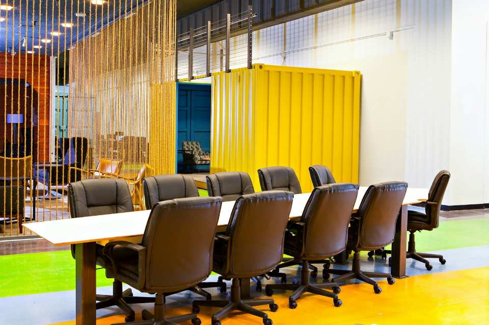Modern office conference room with a long white table and black leather rolling chairs, separated by a rope curtain partition and backed by a bright yellow corrugated shipping container wall partition in a creative coworking space near Mission District, San Francisco (37.767296, -122.419498). This image captures a dynamic and industrial-chic conference room designed for collaborative meetings in a modern San Francisco office setting. The space features a long table surrounded by comfortable black office chairs, set against a backdrop of bold colors, including the striking yellow wall of a repurposed shipping container module. Rope partitions offer semi-privacy, reflecting the innovative and functional design trends popular in the Mission District area.