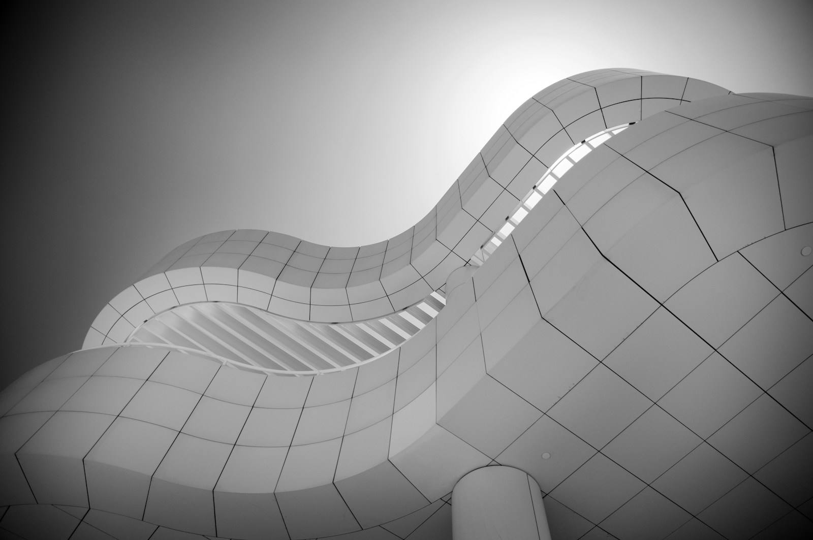 Low-angle black and white view of a highly curved modern building facade with tessellated white panels and diagonal openings, illustrating contemporary structural engineering and abstract design in San Francisco, California. This striking photograph captures the soaring, fluid lines of cutting-edge modern architecture, rendered in stark black and white. The continuous, wave-like surfaces of the exterior panels create a mesmerizing geometric pattern against the bright sky. This abstract interpretation highlights the innovative structural design often celebrated in prominent cultural institutions throughout the San Francisco Bay Area, appealing to those seeking inspirational architectural excellence.