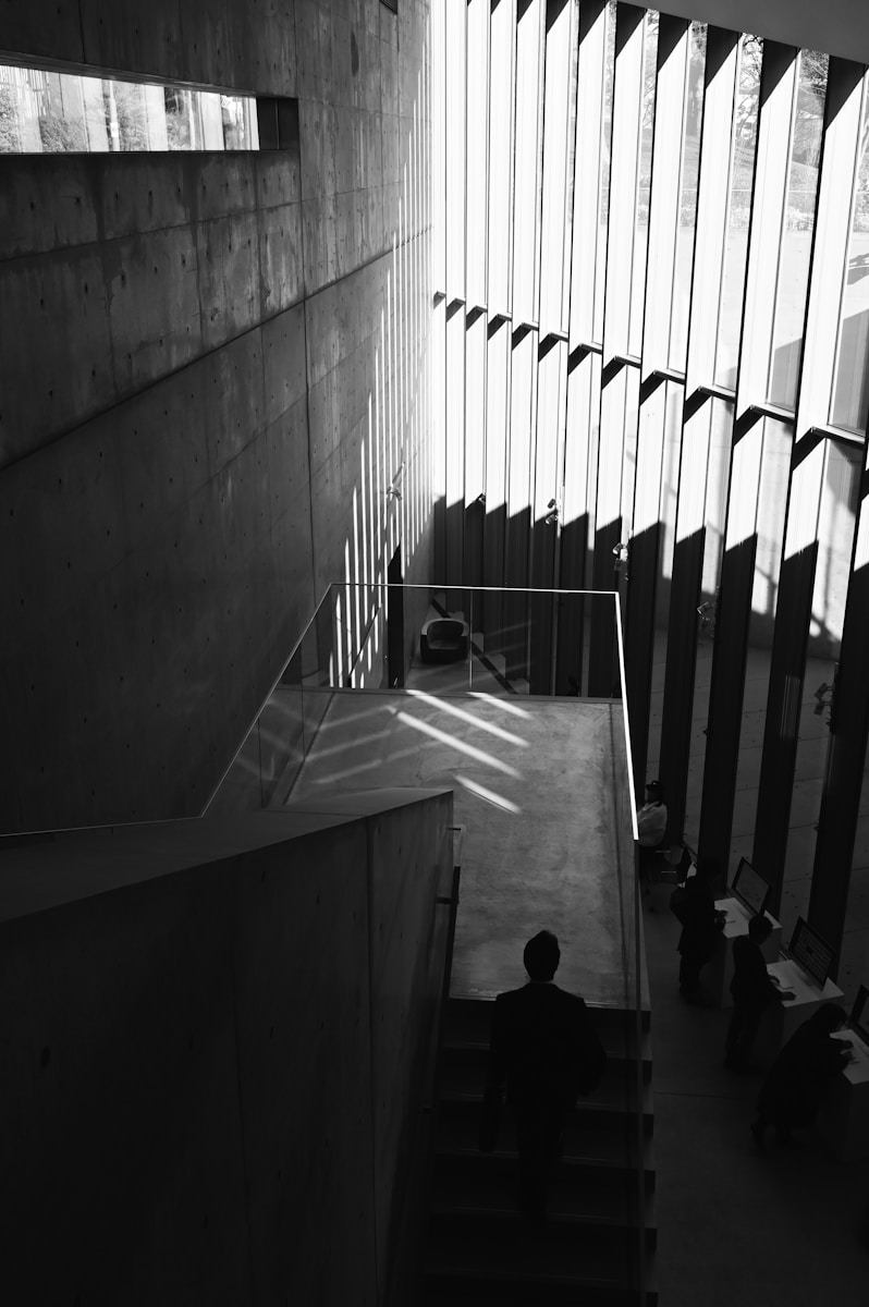 Black and white architectural photograph of a man descending a severe concrete staircase inside a modernist public building in San Francisco, emphasizing dramatic shadows cast by large vertical window fins in the multi-story atrium near Downtown. This striking black and white photo captures the stark beauty of contemporary institutional architecture in San Francisco. The image highlights geometric forms, massive concrete walls, and the dramatic use of natural light filtered through tall vertical windows. A solitary figure navigates the minimalist staircase, underscoring the scale and contemplative atmosphere of the interior space, potentially located near the bustling SOMA district.