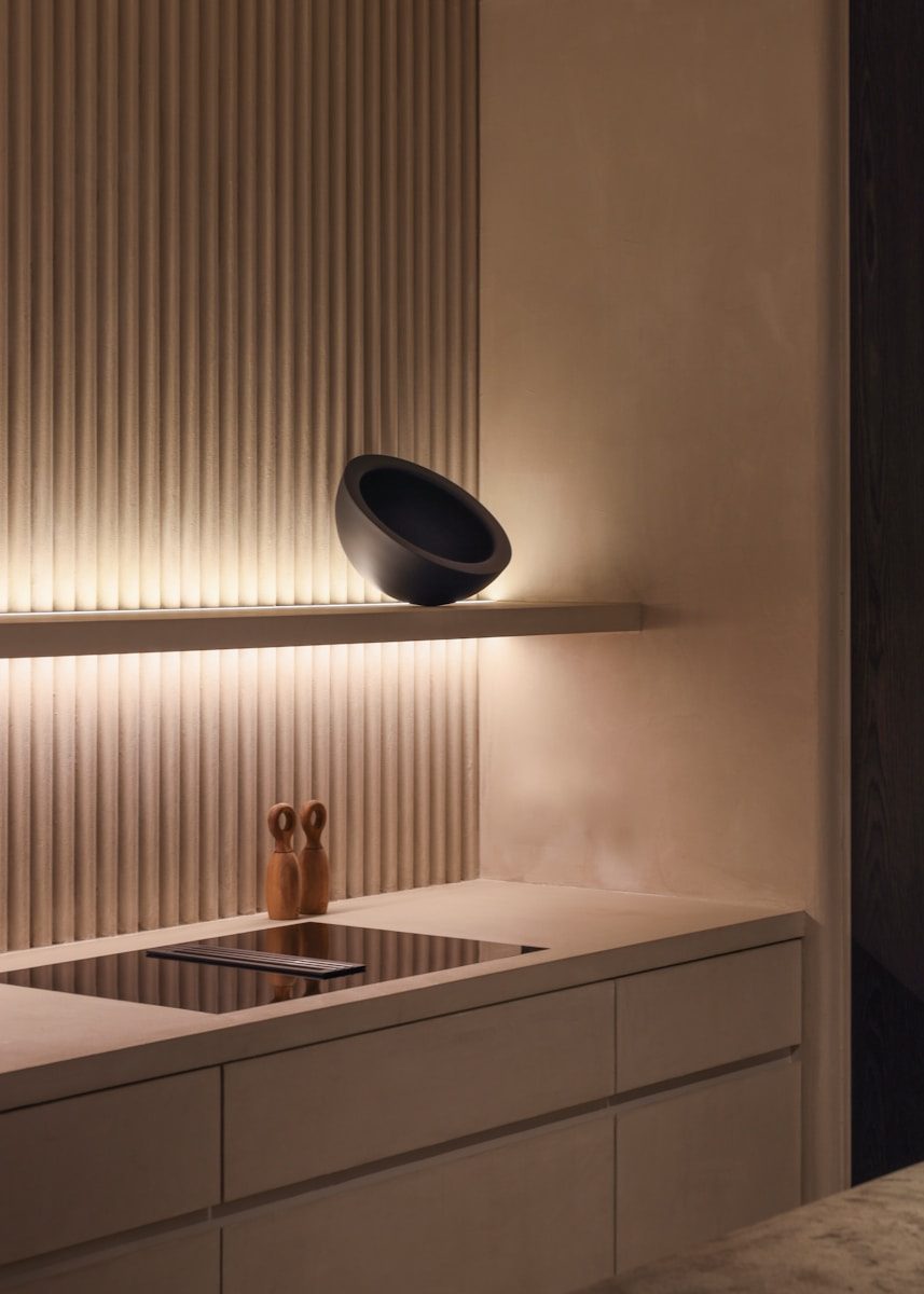 Minimalist modern kitchen design in San Francisco featuring neutral-toned fluted wall paneling, warm LED under-shelf lighting, an integrated induction cooktop, and floating shelf holding dark sculptural decor. This photo captures the essence of luxury minimalist design, perfect for high-end residential projects like those commonly found across San Francisco. The focal point is the subtle texture of the vertical fluted backsplash, beautifully highlighted by warm under-shelf LED strip lighting. Clean lines dominate the entire composition, from the seamless, handleless cabinetry to the integrated induction cooktop, ensuring maximum functionality without sacrificing visual serenity. This sleek design showcases a commitment to refined, contemporary sophistication in modern urban living.
