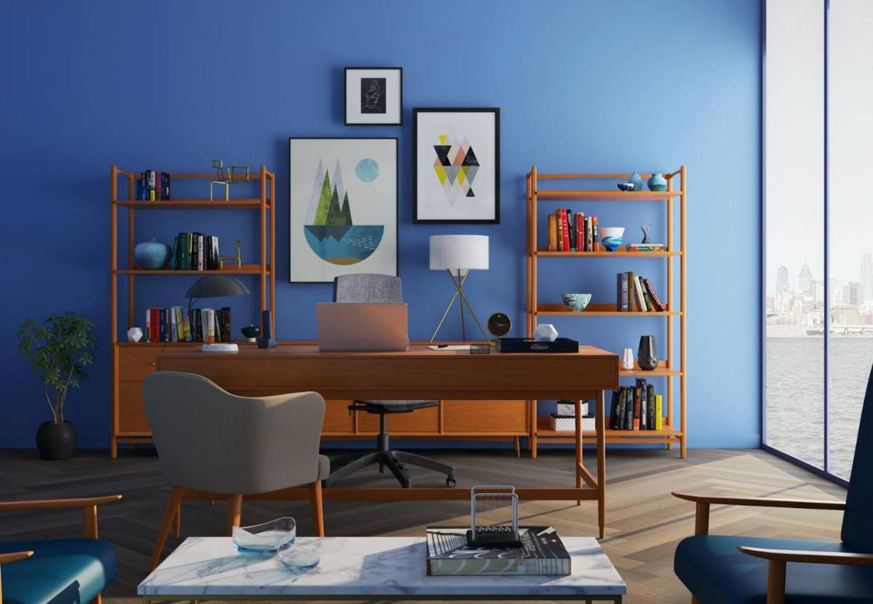 Professional Mid-century modern home office interior in San Francisco featuring bold cobalt blue walls, wooden bookshelves stocked with books, a large teak wood desk with a laptop, and a panoramic window overlooking the city skyline across the water. This sophisticated interior features a vibrant cobalt blue wall backdrop perfectly complementing the warm tones of the Mid-century modern wood desk and shelving units. Designed as a productive and aesthetically pleasing workspace, the room boasts excellent natural light and a stunning cityscape view across the bay near the San Francisco core. Accessories like abstract art prints, a white marble coffee table, and comfortable blue and gray seating complete this high-end residential office design.