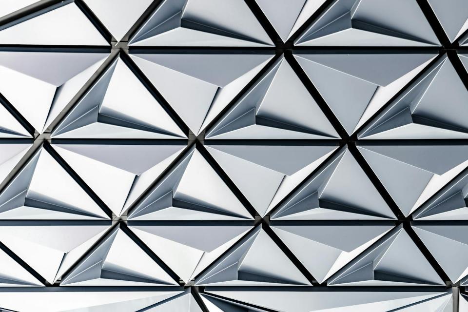 Abstract view of a modern geometric facade featuring repeating triangular metal panels in light gray and white, showcasing innovative architectural cladding design on a structure located near the Mission District of San Francisco (Lat 37.767, Lng -122.419). This image captures the intricate detail of a contemporary architectural surface, characterized by its sharp, tessellated pattern of triangular elements. The interplay of light and shadow across the metallic gray panels highlights the depth and complexity of the design. This modern facade detail is representative of cutting-edge building materials and innovative aesthetics found in new developments across the vibrant Mission District of San Francisco, appealing to architects and design enthusiasts seeking structural inspiration.