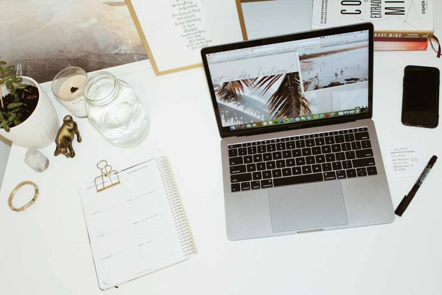 Overhead view of a modern and organized remote desk setup featuring a MacBook Pro, spiral planner, smartphone, potted succulent, and inspirational items, symbolizing productive work in the San Francisco Bay Area. This overhead shot captures an aesthetically pleasing and highly organized remote workspace, ideal for entrepreneurs or freelancers operating in the competitive San Francisco market. The clean setup features a silver MacBook Pro, a detailed weekly planner, and mindfulness elements like plants and crystals, promoting focused creativity and work-life balance necessary for Bay Area professionals thriving near the 37.76 latitude zone.